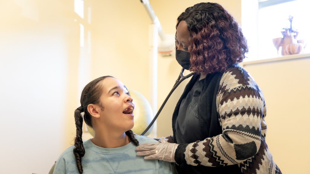 In bright room painted tan, a young girl with black pigtails smiles and looks up at a nurse practitioner as the NP holds a stethoscope to her back at Woods Healthcare's outpatient Medical Center. 