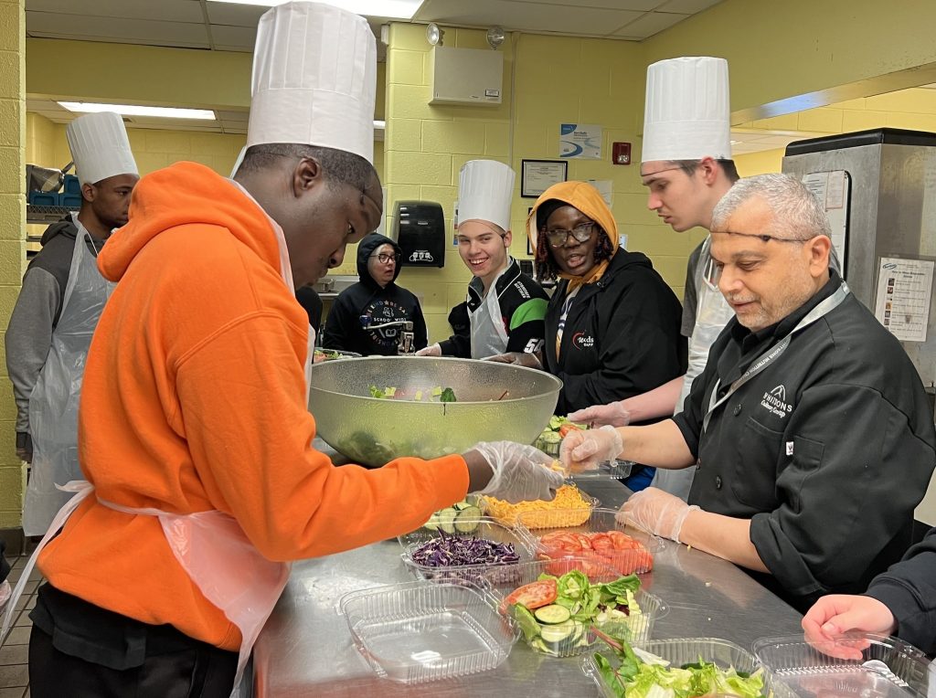 student in orange hoodie is making a salad with advice from a man wearing a black chef shirt